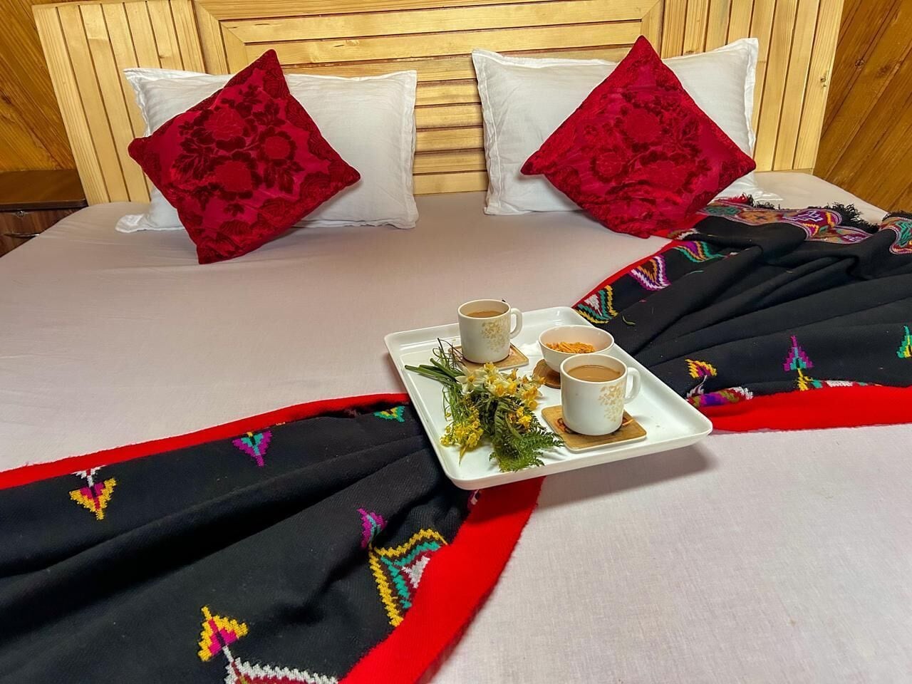 A neatly made bed with a tray of coffee and flowers, red pillows, and a black and red embroidered blanket.