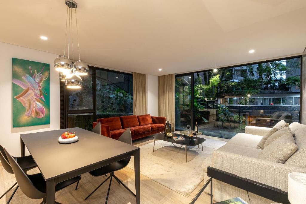 Een moderne woonkamer met een eetgedeelte, een rode bank, een grijze bank en een groot raam met uitzicht op groen.