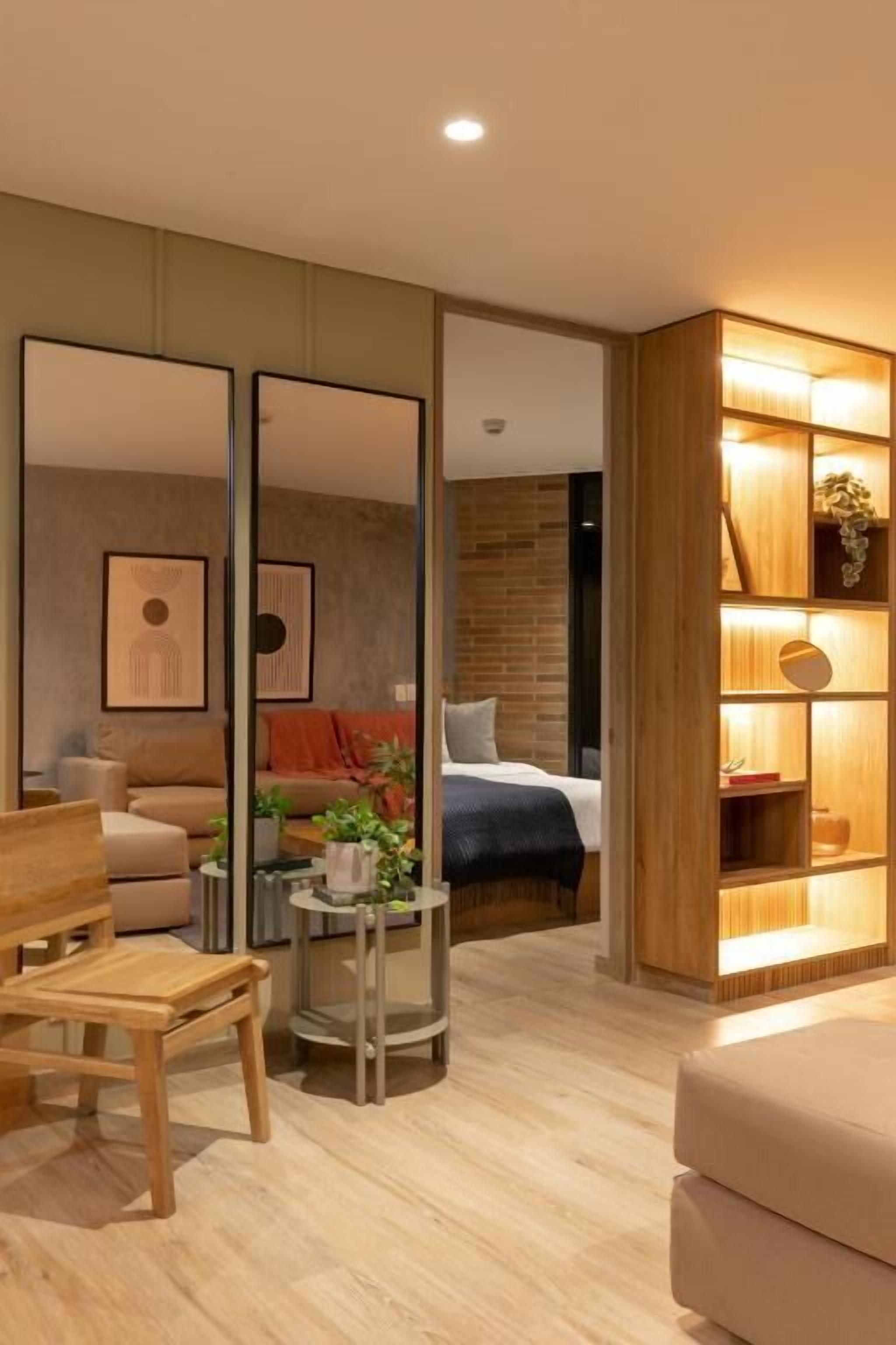 Una camera d'albergo moderna con pavimento in legno, un letto con coperta blu, un comodino in legno con una pianta, una poltrona e una libreria in legno illuminata da luci calde.