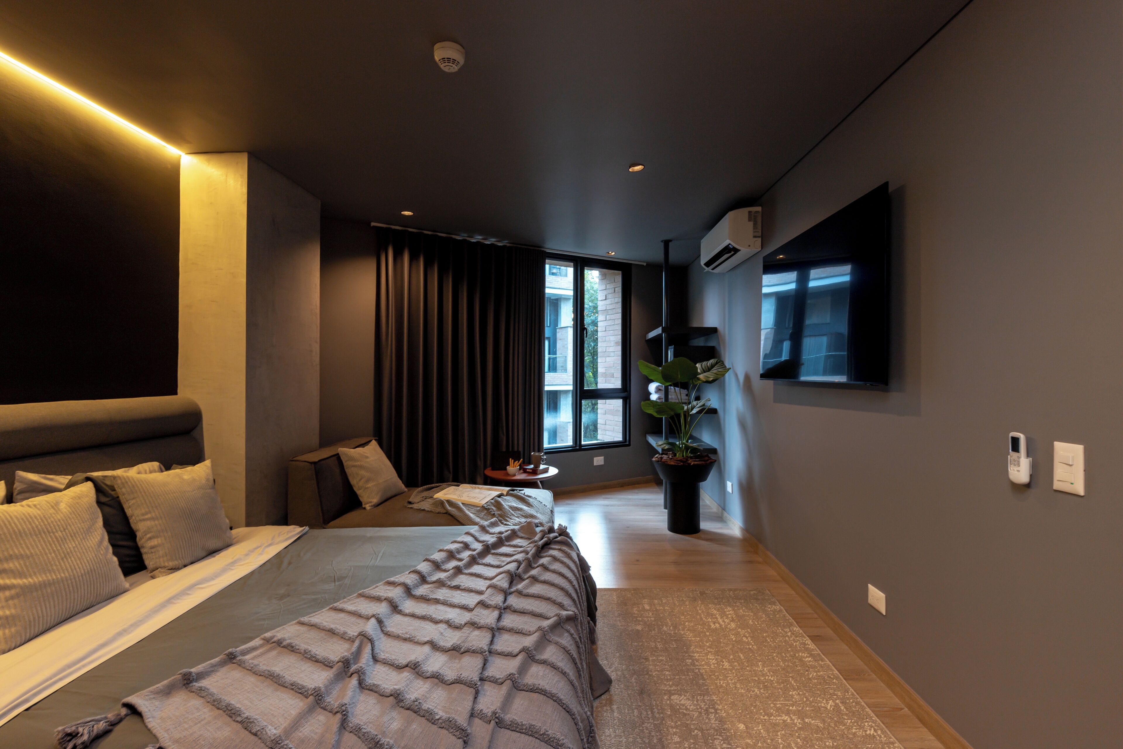 A modern bedroom with a large bed, a bedside table, a potted plant, and a window with curtains.