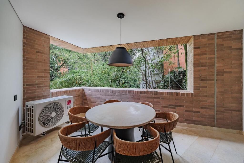 Een moderne eetruimte met een ronde tafel en rieten stoelen, een airconditioning en uitzicht op groen door een groot raam.