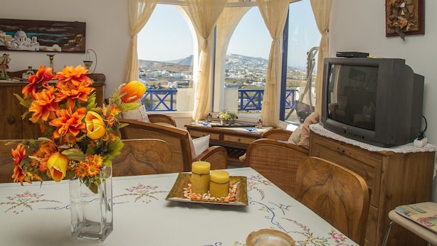 A dining area with a table set for two, a vase of flowers, and a view of the city through large windows. - Villa Eva with Panoramic view (Santorini)