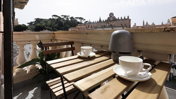 A wooden table with two white cups and saucers, a bench, and a view of historic buildings and trees. - Plaza Suites Rome (Rome)
