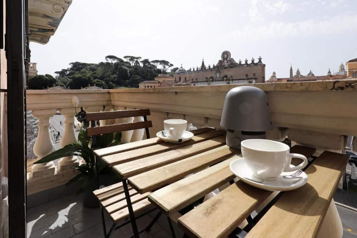 A wooden table with two white cups and saucers, a bench, and a view of historic buildings and trees.