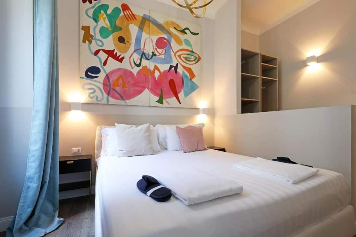 A hotel room with a large bed, white bedding, a colorful abstract painting on the wall, and a built-in shelf unit.
