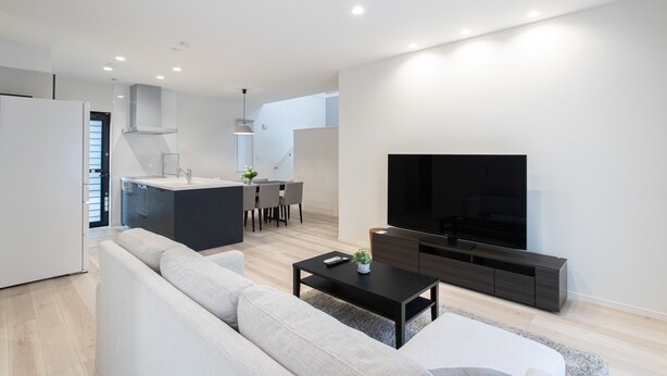 A modern living room with a sofa, coffee table, TV, and kitchen area in the background. - Mova Villa Okino (Kikuyo)