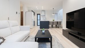 A modern living room with a sofa, a coffee table, and a TV. There is a kitchen area in the background with a dining table and chairs. - Mova Villa Okino (Kikuyo)