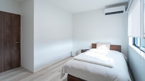 A bedroom with a bed, a nightstand, a wall-mounted air conditioner, and a window with blinds. - Mova Villa Okino (Kikuyo)