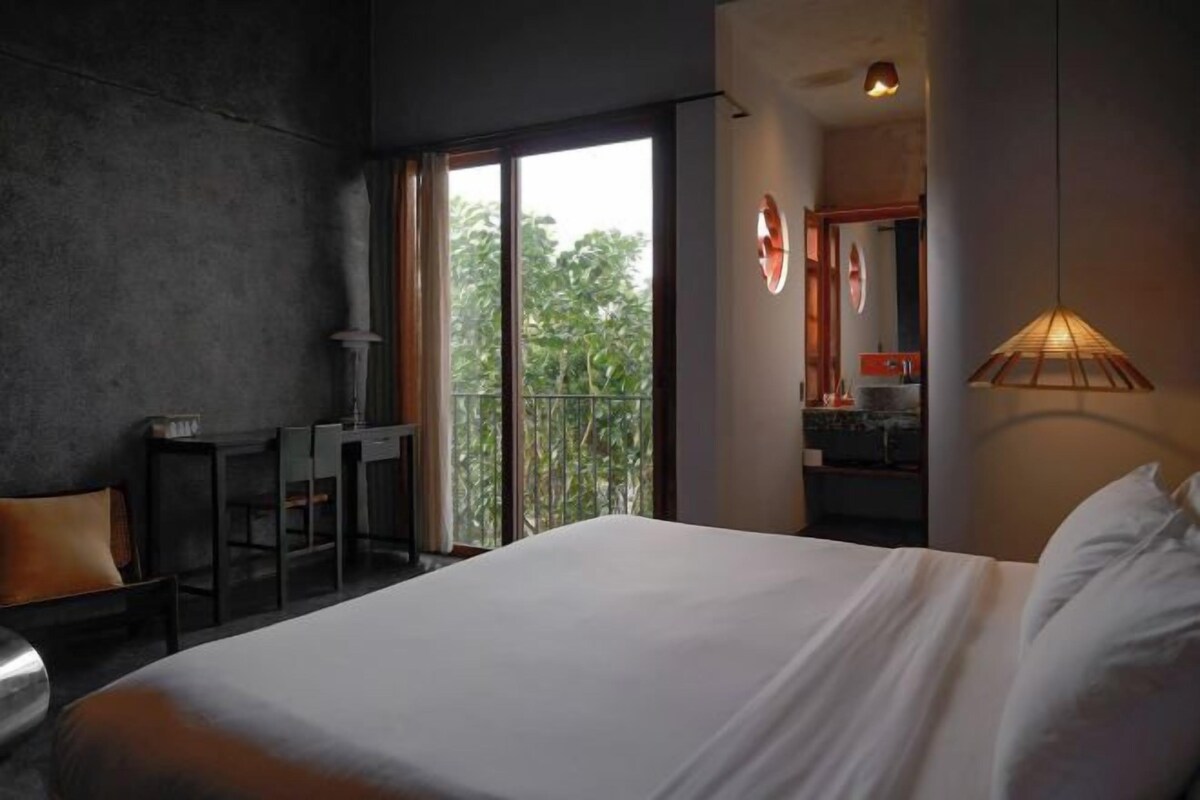 A hotel room with a bed, a desk, and a balcony with a view of trees.