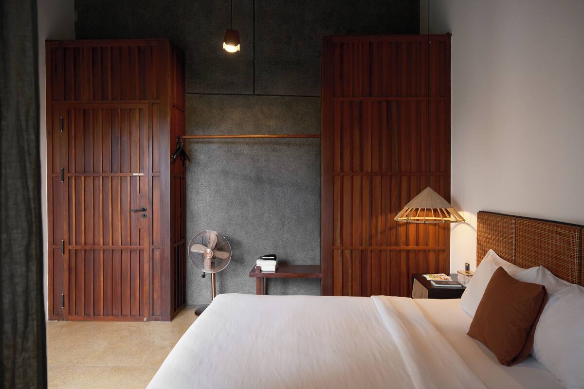 A bedroom with a bed, wooden wardrobe, a nightstand, a lamp, and a fan.