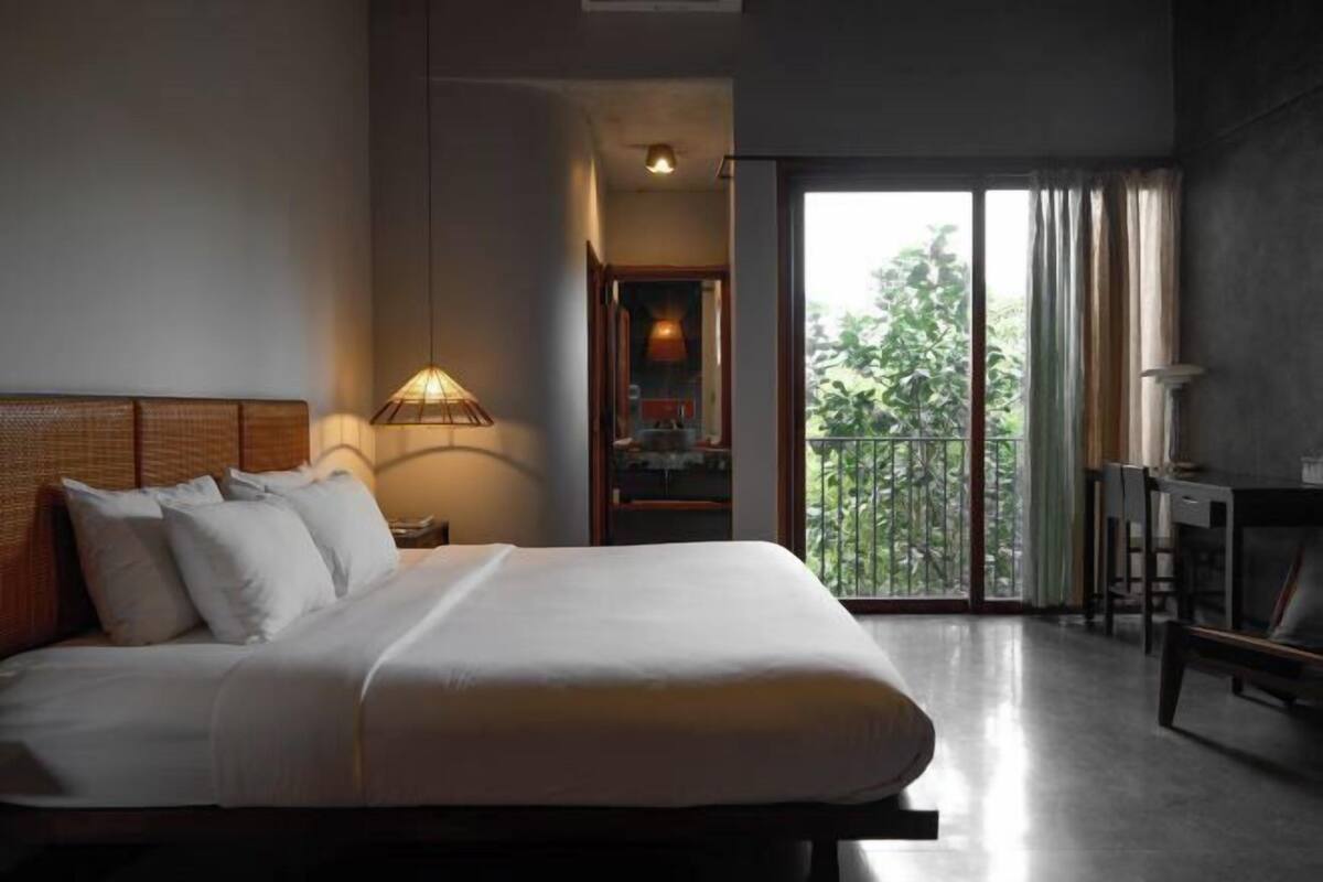 A hotel room with a bed, a desk, a lamp, and a view of greenery through the window.