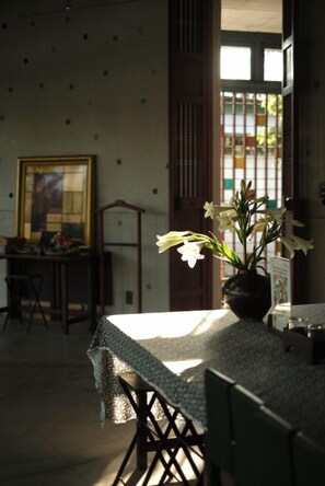 A dining area with a table covered by a patterned tablecloth, a vase with white flowers, and a painting on the wall. - De Hué Space (Hue)