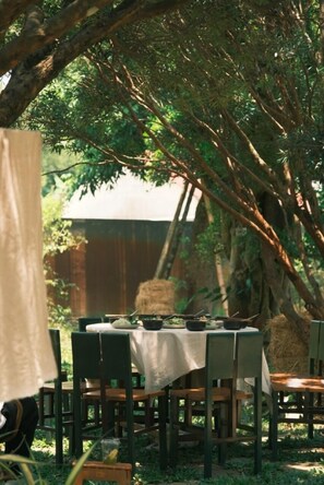 An outdoor dining area with a table set under a tree, surrounded by chairs. - De Hué Space (Hue)