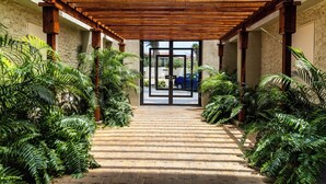 A covered walkway with a wooden ceiling, flanked by lush greenery and leading to a glass door.