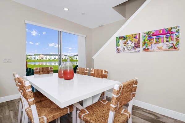 A dining area with a white table and wicker chairs, a large window with a view of houses, and two colorful paintings on the wall.