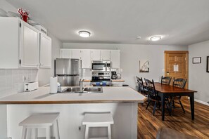 Uma cozinha moderna com armários brancos, uma ilha central com banquetas, uma mesa de jantar com cadeiras e piso de madeira.