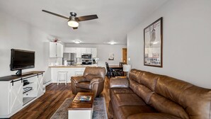 A modern living room with a brown leather sofa, a wooden coffee table, and a TV on a wooden stand. There is a ceiling fan, a kitchen in the background, and a framed picture on the wall. - Hike In/Hike Out w/Slope Views + 3 Hot Tubs-Ski/Town Shuttle-Sauna-Gym-Gas Fireplace-Deck-Updated (Steamboat Springs)