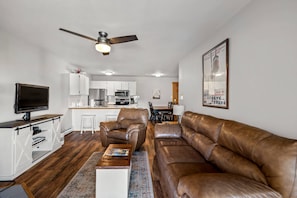 Uma sala de estar moderna com um sofá de couro marrom, uma mesa de centro de madeira e uma TV em um móvel de madeira. Há um ventilador de teto, uma cozinha ao fundo e um quadro na parede.