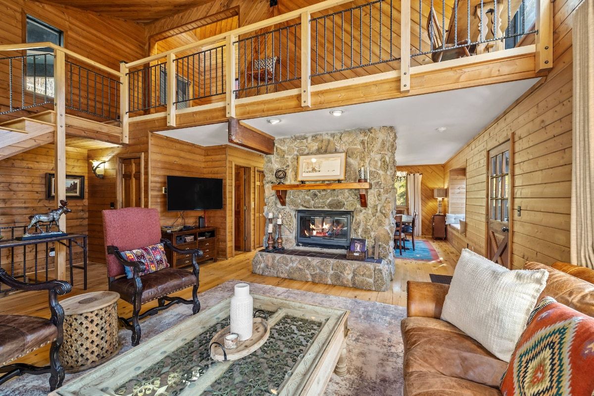 A spacious log cabin interior with a stone fireplace, wooden furniture, and a balcony.