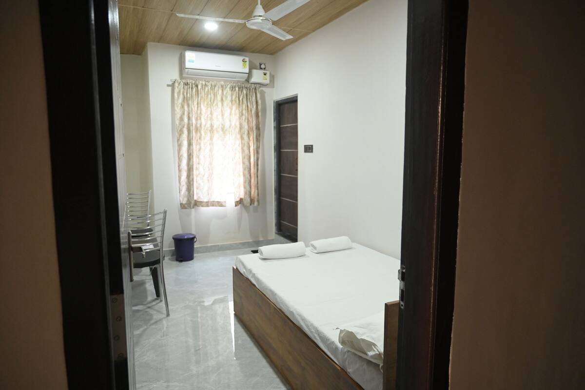 Une chambre comprenant un lit, un ventilateur de plafond, un appareil de climatisation, une fenêtre avec des rideaux et une petite table avec une chaise.