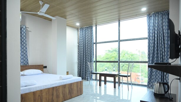 Quarto de hotel moderno com cama de madeira, escrivaninha com chaleira e uma janela grande com cortinas.