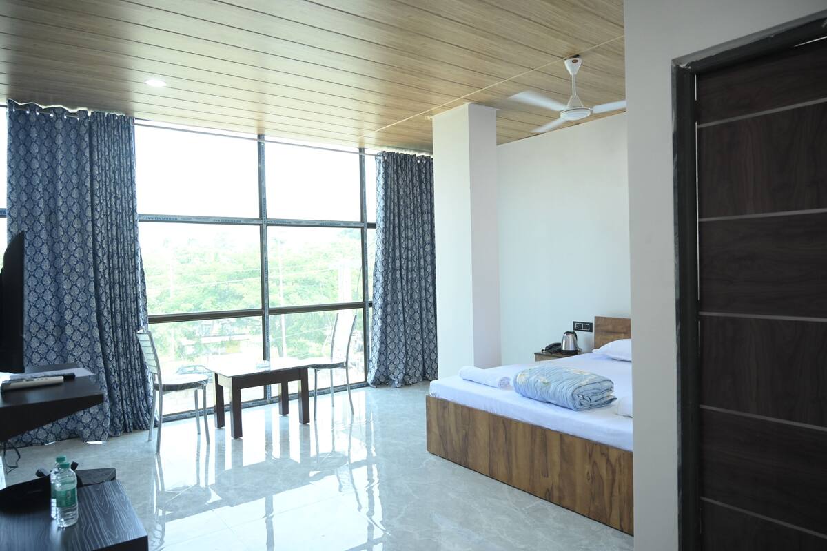 Une chambre d’hôtel moderne avec un plafond en bois, une grande fenêtre avec des rideaux, un lit avec une tête de lit en bois, une table de chevet et une chaise.
