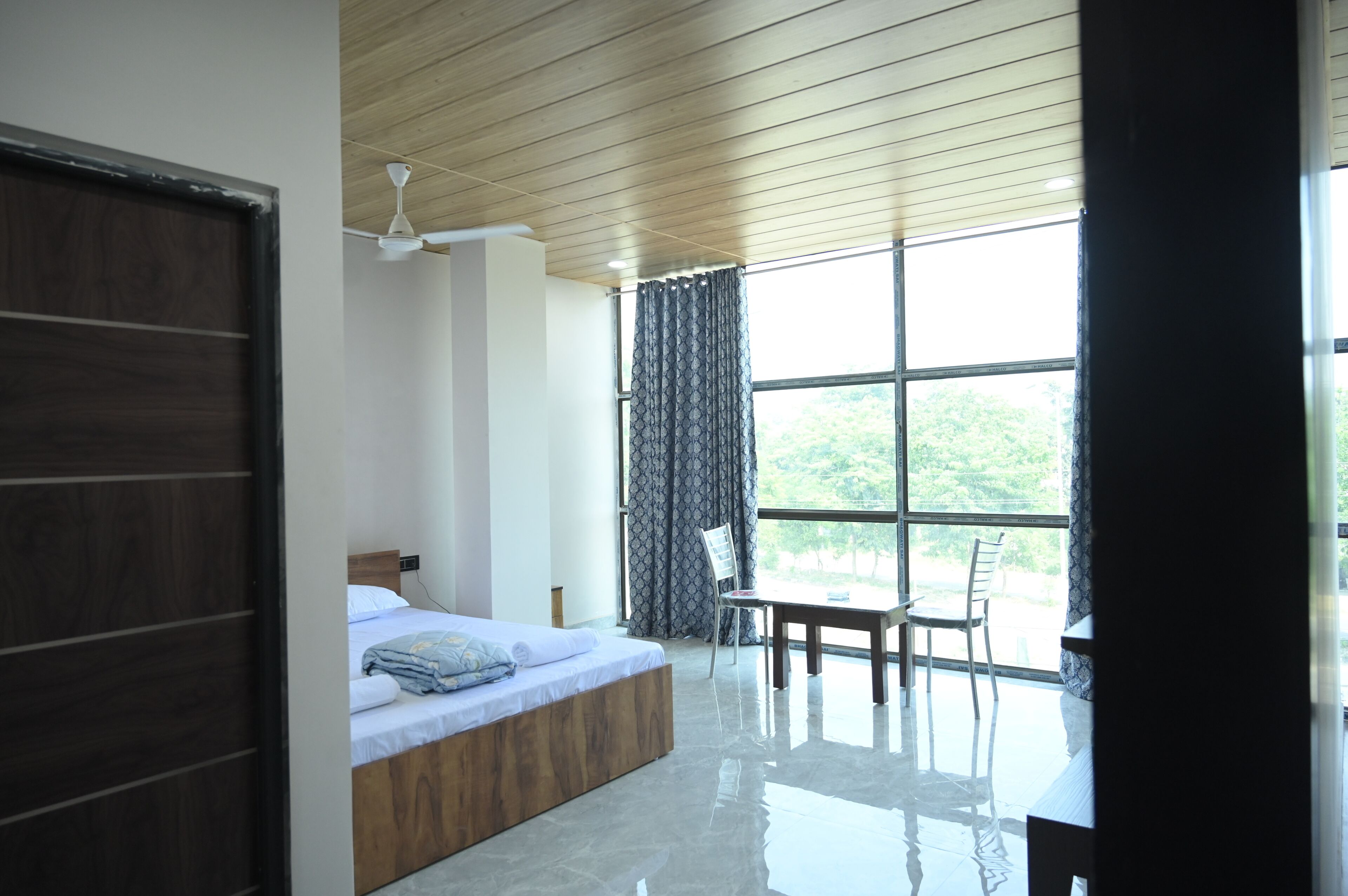 A modern bedroom with a wooden ceiling, a large window with curtains, a bed, a table, and a chair.