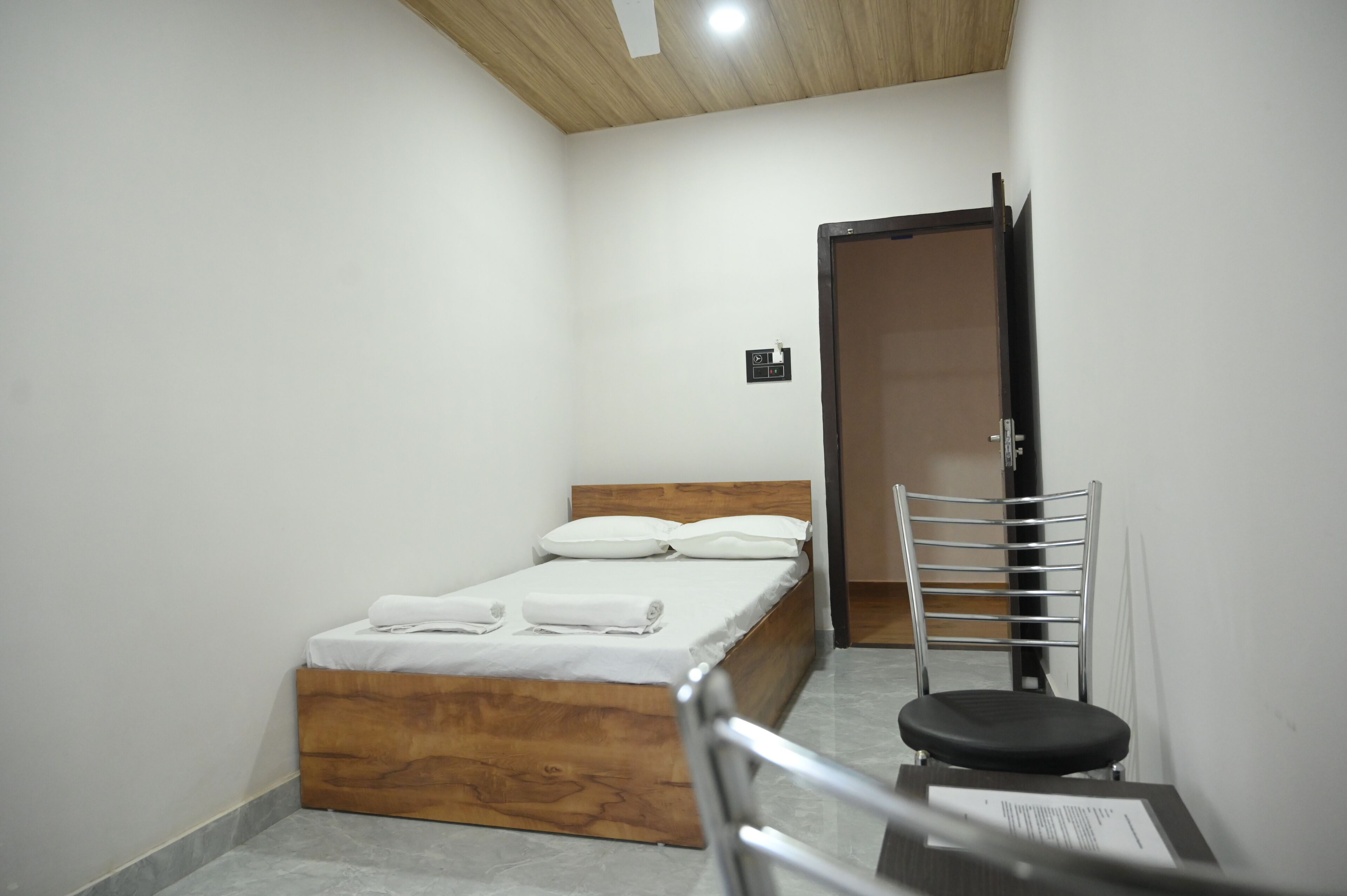 Ένα μικρό, καθαρό δωμάτιο με ένα ξύλινο κρεβάτι, μια καρέκλα και μια πόρτα που οδηγεί σε ένα άλλο δωμάτιο.