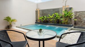 Zona de piscina exterior moderna con mesa de vidrio y dos sillas, rodeada de plantas en macetas y un muro de piedra.