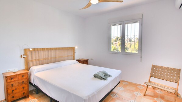 Une chambre à coucher avec un lit, des tables de chevet en bois, un banc, une fenêtre donnant sur un beau paysage et un ventilateur de plafond.
