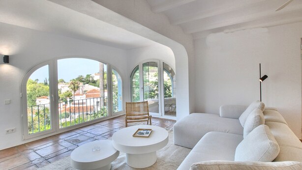A spacious living room with a large white sofa, a round coffee table, and a wicker chair. There are large arched windows providing a view of the outside. - Luxury villa with sauna and private pool in Denia 10P (Denia)