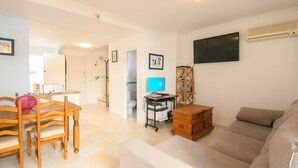 Uma sala de estar com sofá, mesa de madeira com cadeiras, televisão fixada na parede e um pequeno armário de madeira.