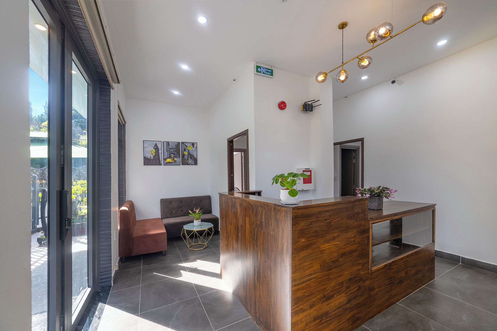 Una moderna zona de recepción con un mostrador de madera, un sofá, una mesita redonda con una planta y un ventanal con vistas al exterior.
