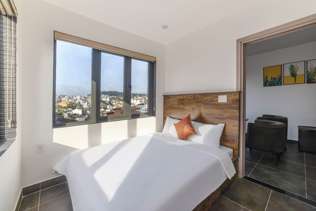 Ein modernes Hotelzimmer mit einem großen Bett, einem Holzkopfteil, Blick auf die Stadt und einer kleinen Sitzecke mit zwei Stühlen.