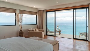 Quarto com uma cama grande, um sofá, vista para o mar através de janelas do chão ao teto, e um vaso grande com plantas secas.
