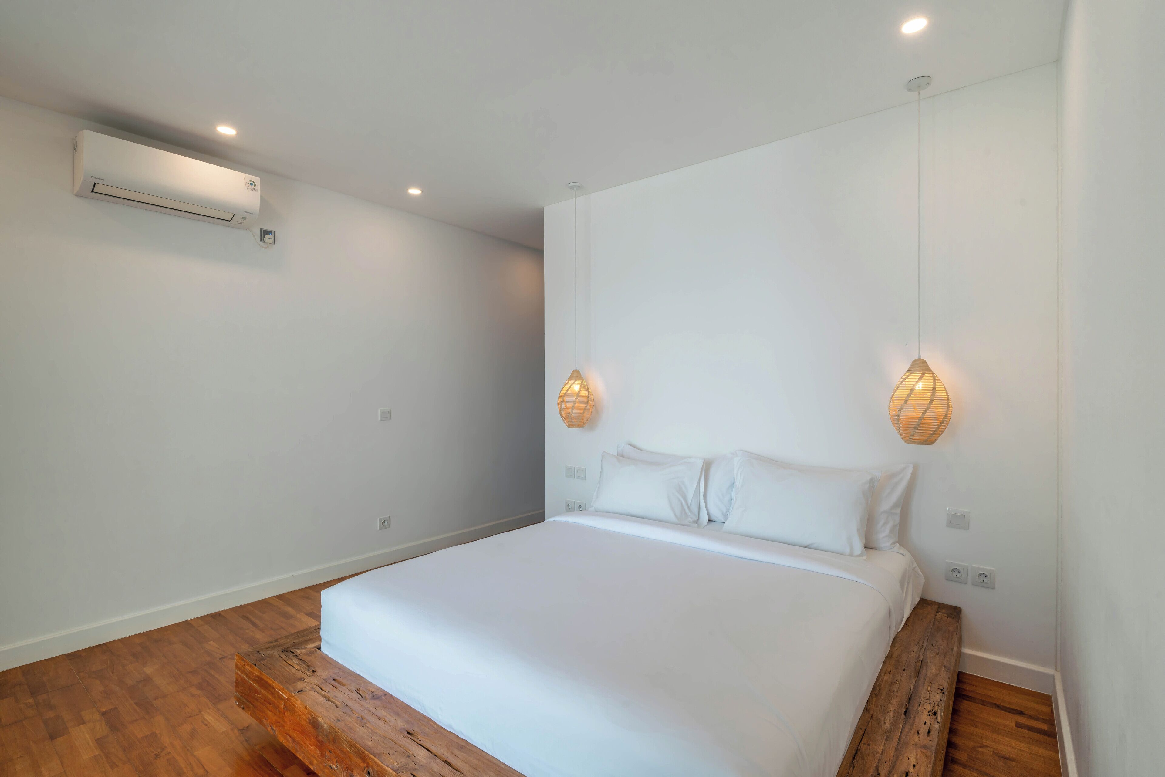 Un dormitorio con piso de madera, una cama blanca, un equipo de aire acondicionado y dos lámparas colgantes.