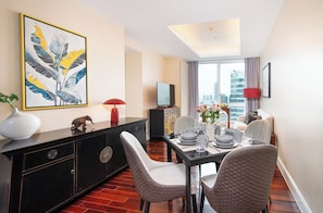 A modern hotel room with a dining area, a large window with a city view, a flat-screen TV, and a decorative plant.