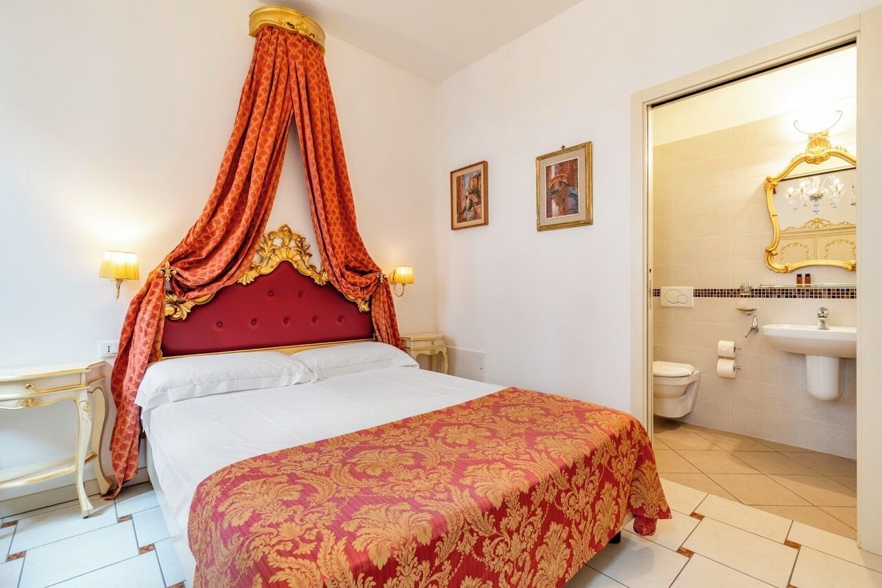Camera d'albergo con un letto a baldacchino, un baldacchino rosso, due lampade da comodino, una testiera imbottita rossa, un coprimaterasso bianco, un piccolo comodino e un bagno con lavandino e water.