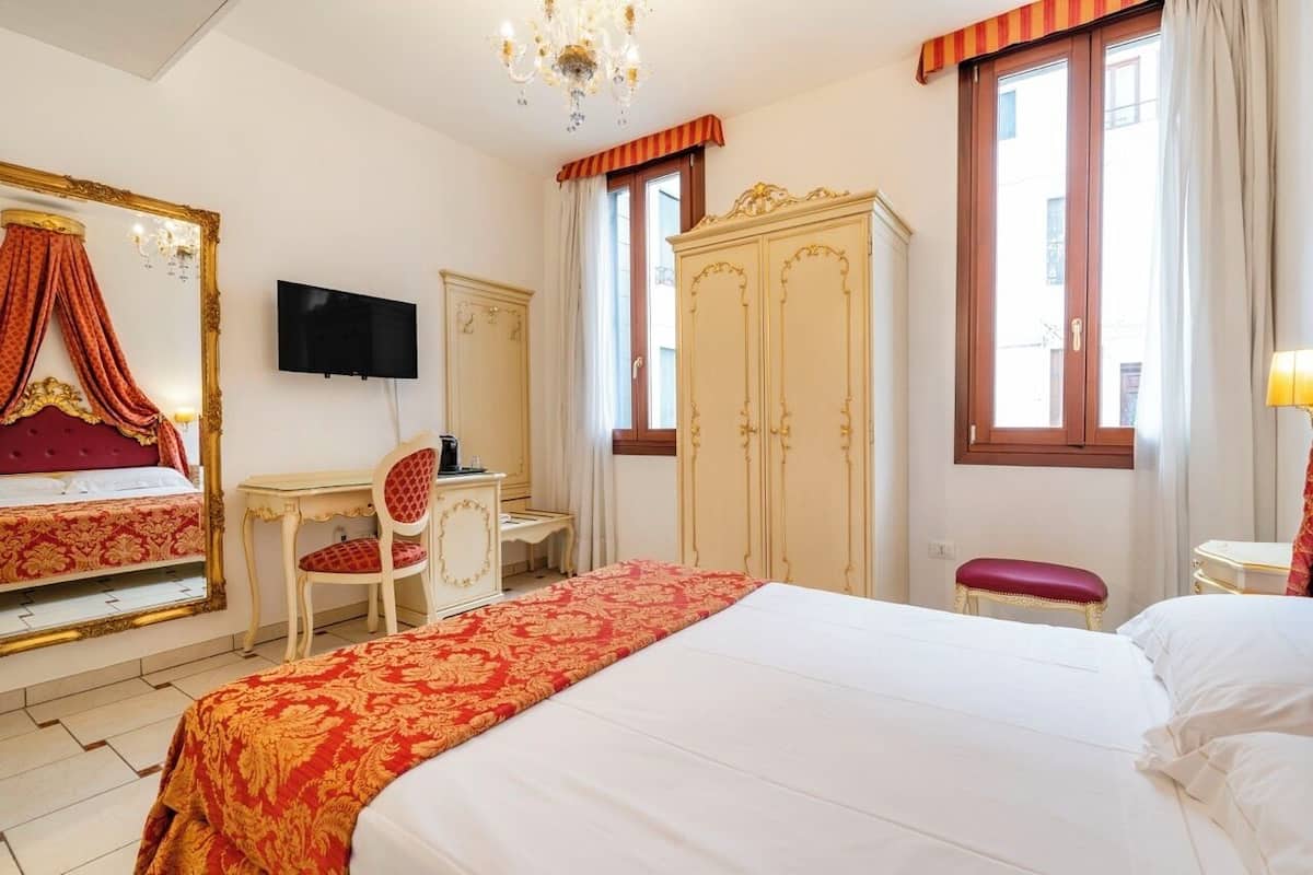 Habitación de hotel con una cama grande, un escritorio, una silla, un armario, televisión y ventana con cortinas.