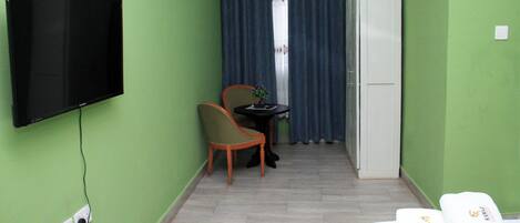 A hotel room with a flat-screen TV mounted on the wall, a small table with a chair, a bed with a green bedspread, and a white wardrobe.