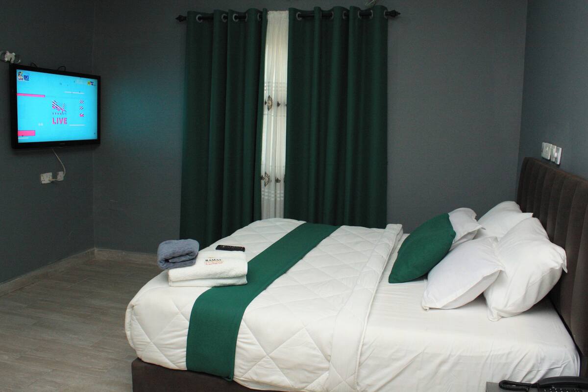 Een net opgemaakt bed met een wit-groene spreideken, een afstandsbediening en een opgevouwen handdoek aan de zijkant.