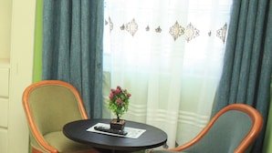 Een kamer met twee houten stoelen rond een klein, rond tafeltje, een plant in een pot op het tafeltje, en een raam met gordijnen.