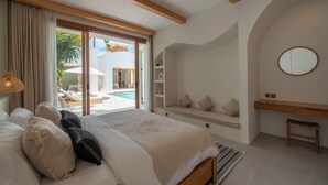 A bedroom with a large bed, a built-in bench, a small table, and a view of an outdoor pool area. - Villa Milana (Canggu)