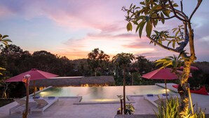 A pool area with lounge chairs and umbrellas, surrounded by tropical plants and a sunset view. - Wijaya Cottage Nusa Penida (Penida Island)