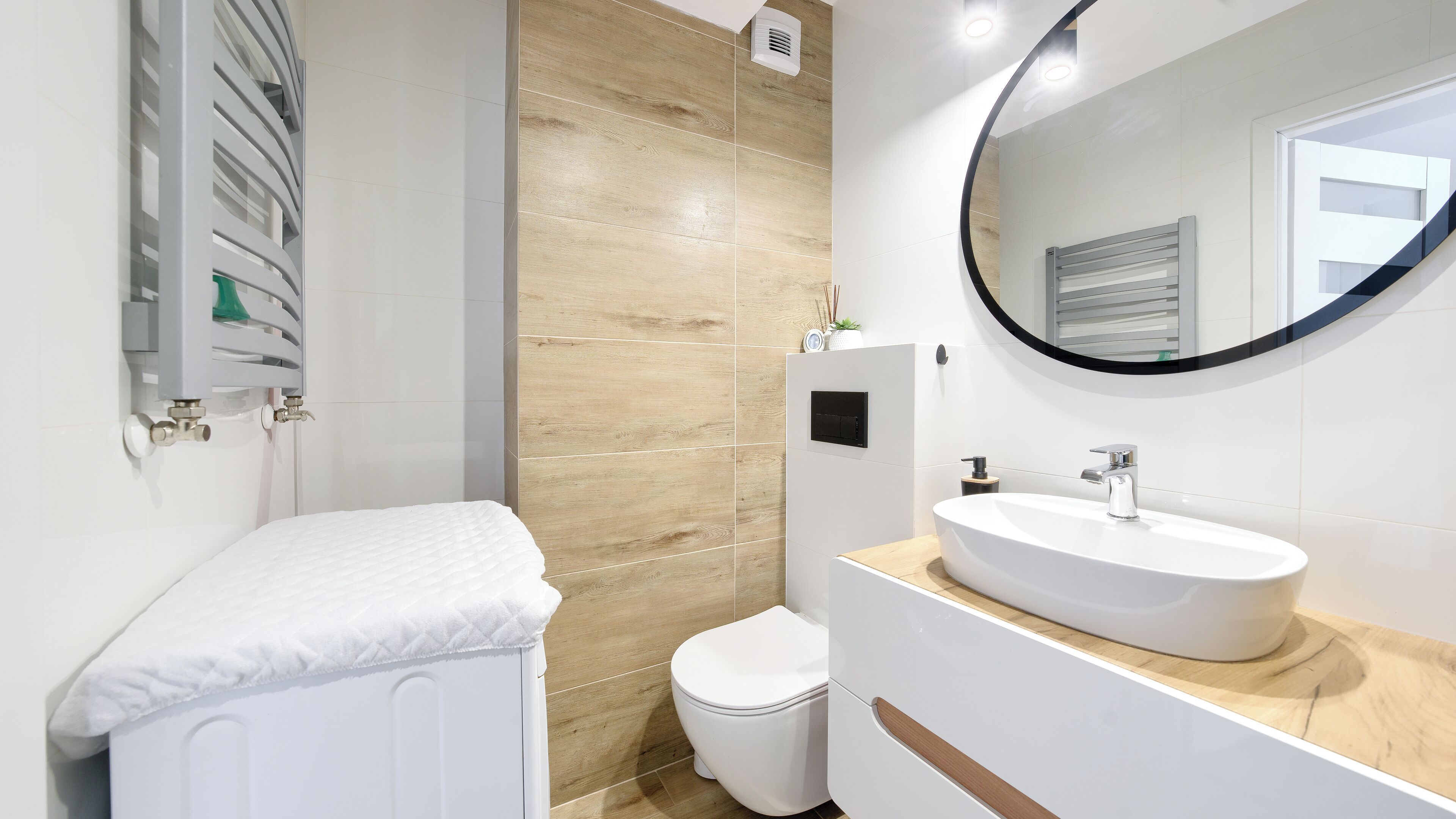 Une salle de bain moderne avec des toilettes blanches, un miroir rond et un lavabo blanc posé sur un plan de travail en bois.