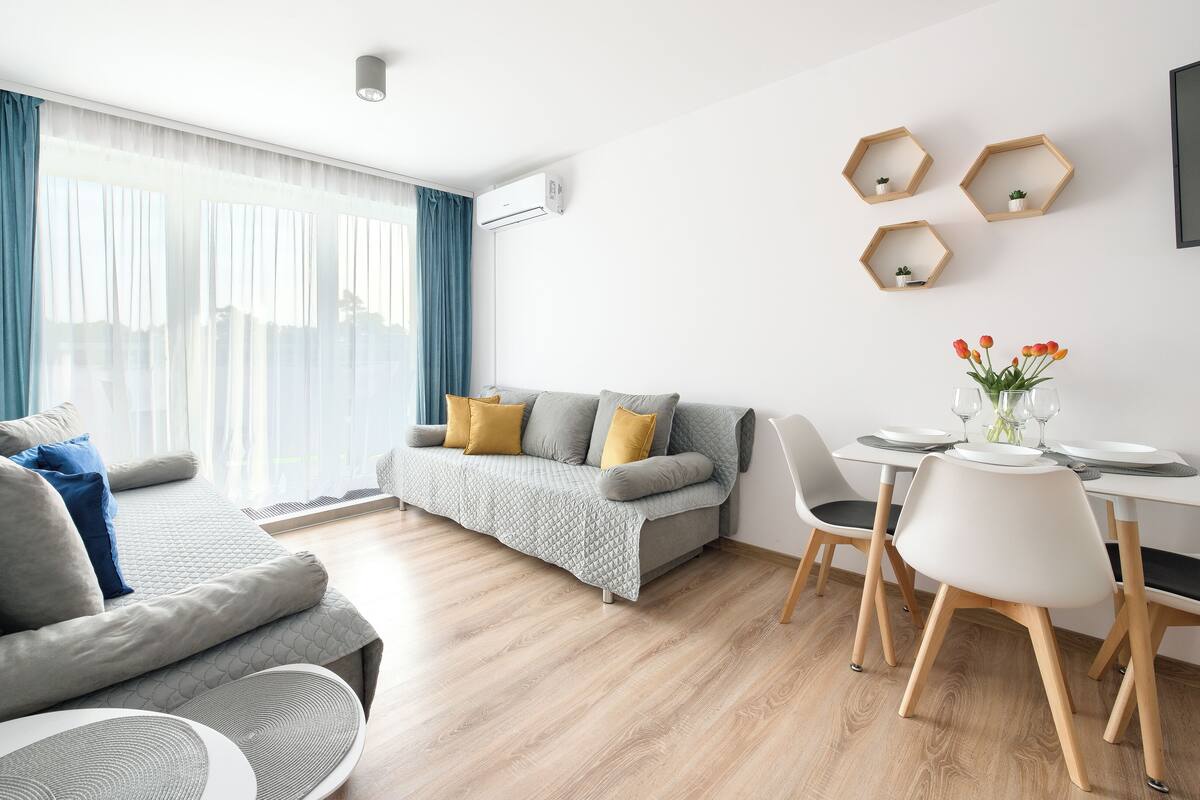 A modern living room with a grey sofa, a dining table with chairs, and a large window with curtains.