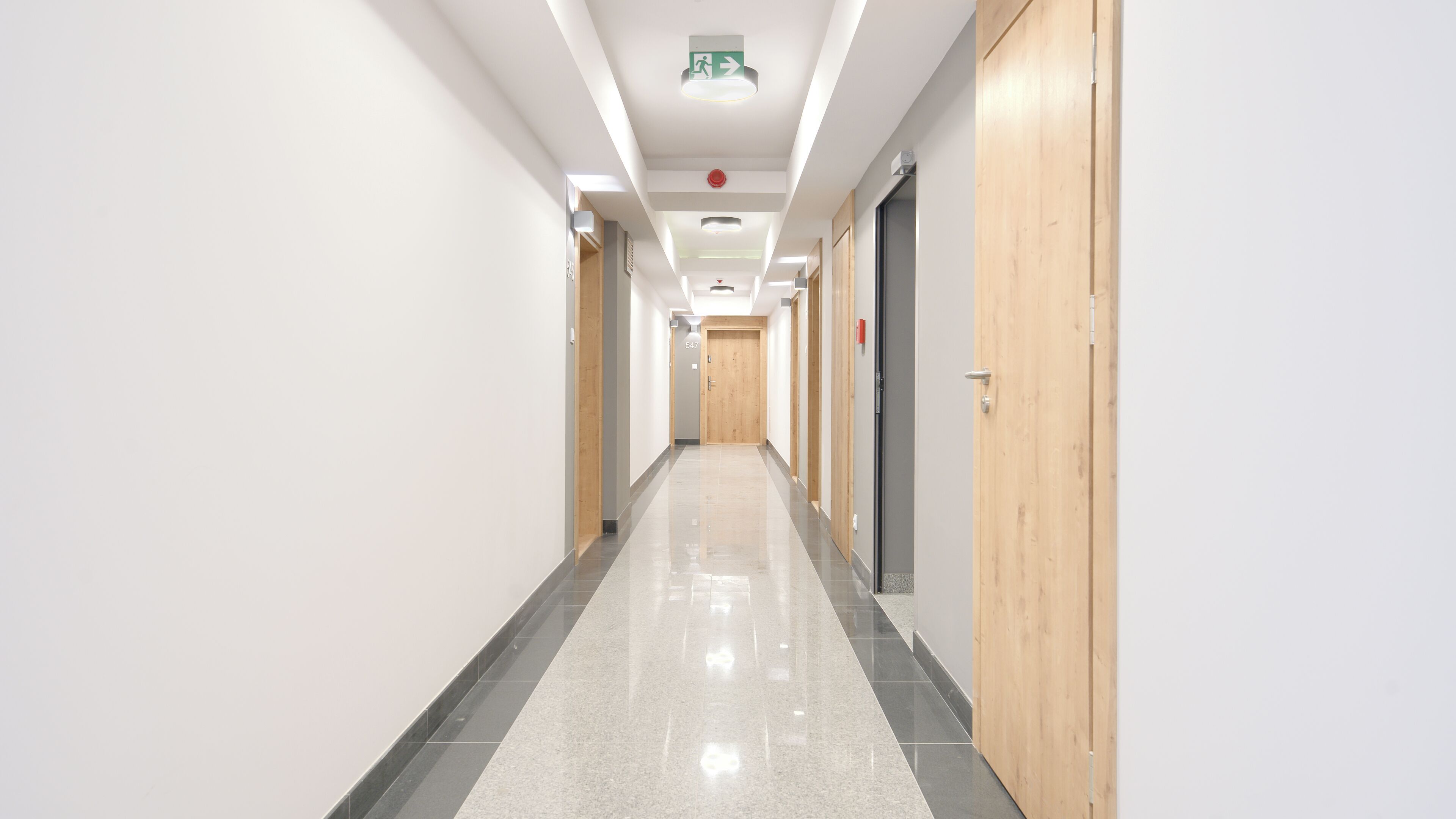 Ein langer, schmaler Korridor mit poliertem Boden, Holztüren und einer Decke mit eingelassener Beleuchtung sowie einem Notausgangsschild.