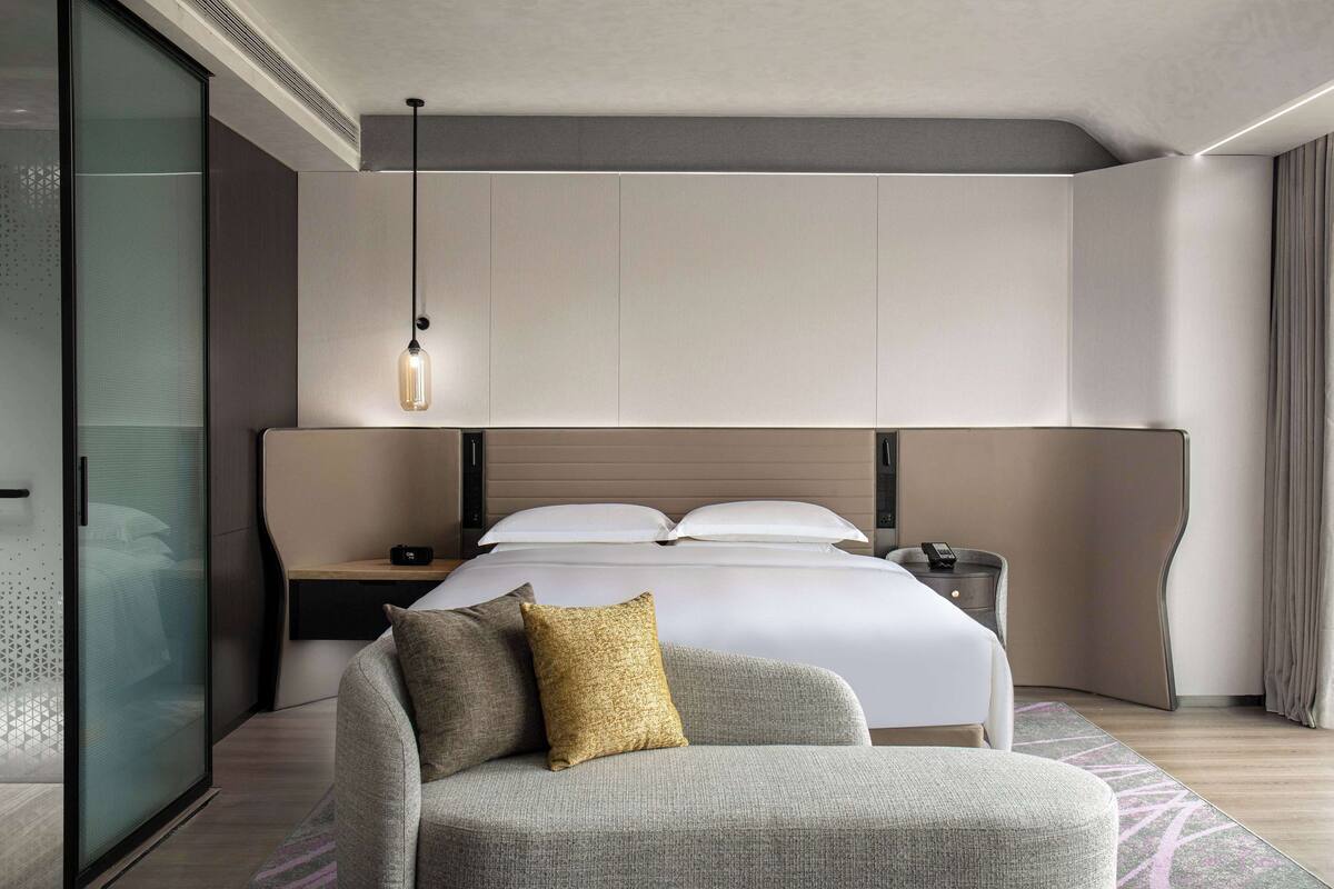 A modern hotel room with a large bed, a bedside table, a lamp, and a sofa.