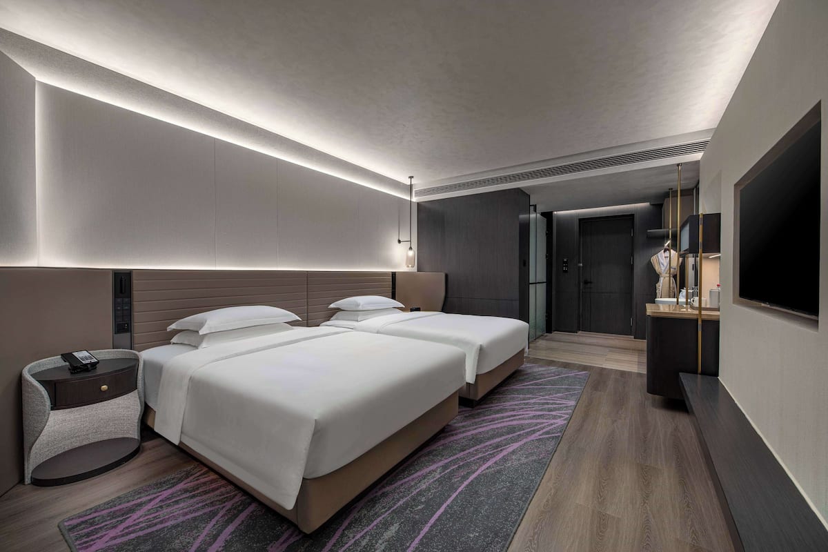 A modern hotel room with two beds, a bedside table with a phone, a TV, and a small seating area with a table and chairs.