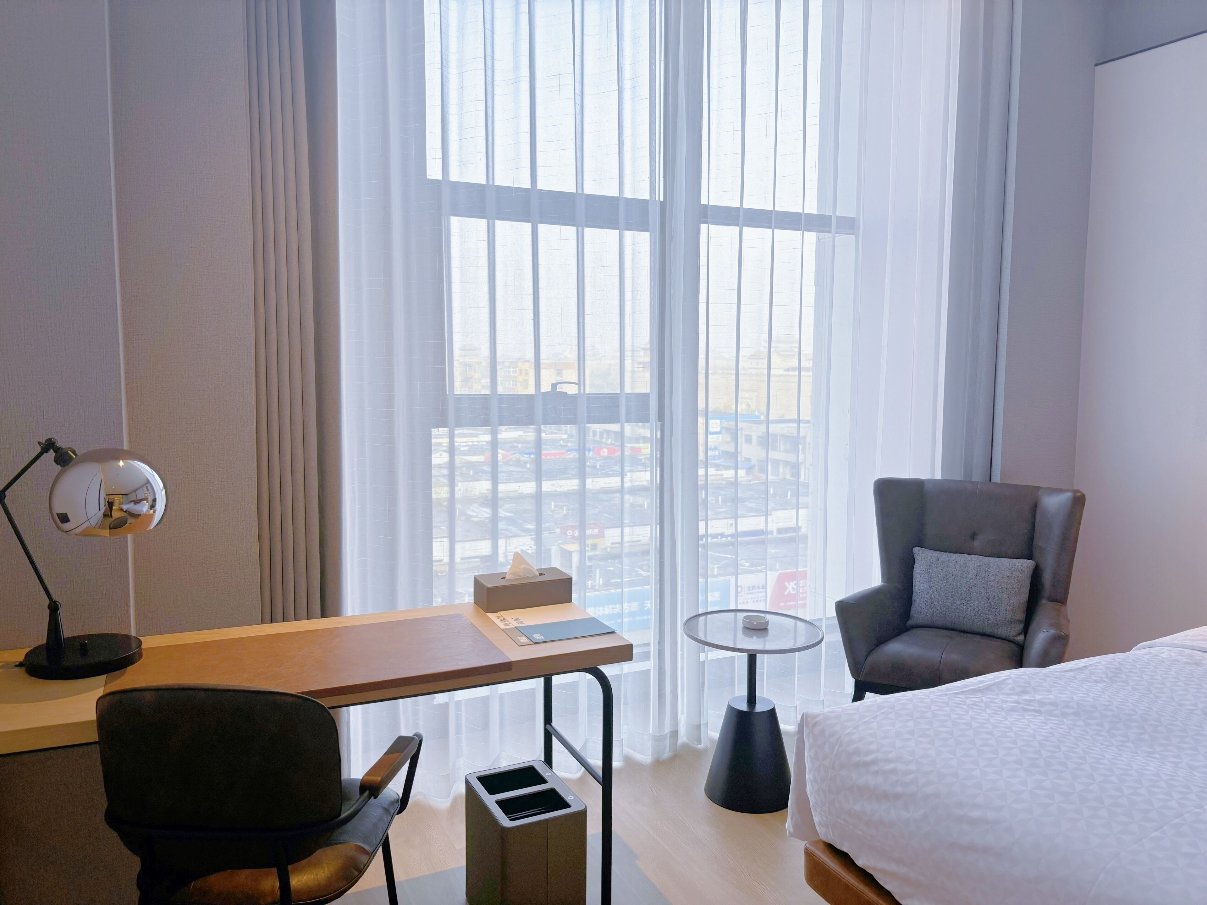 Une chambre d’hôtel avec un lit, un bureau avec une lampe, une chaise, une petite table et une fenêtre donnant sur des immeubles.